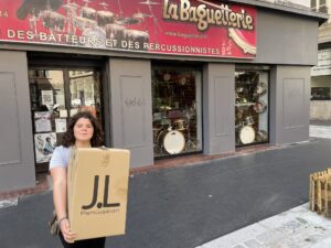 Lilian shows off the box with their new cajon in it in front of "La Baguetterie"
