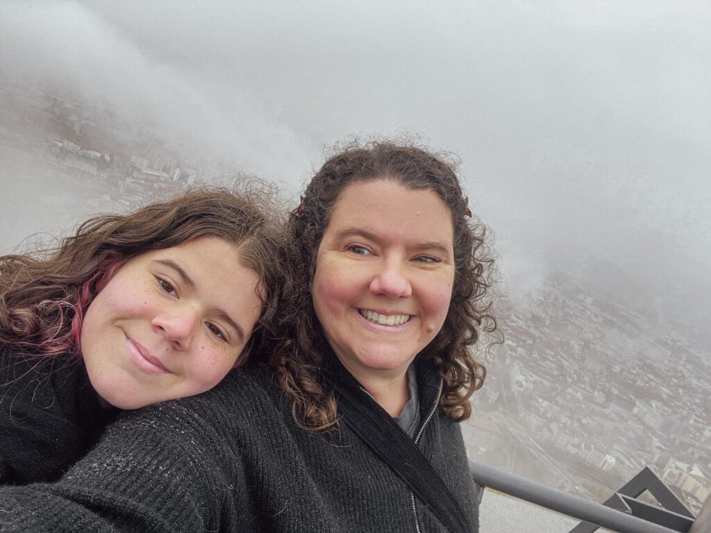 Two smiling women pose together on a foggy day, with a scenic view of a city in the background. They are wearing warm clothing, suggesting a cool climate.