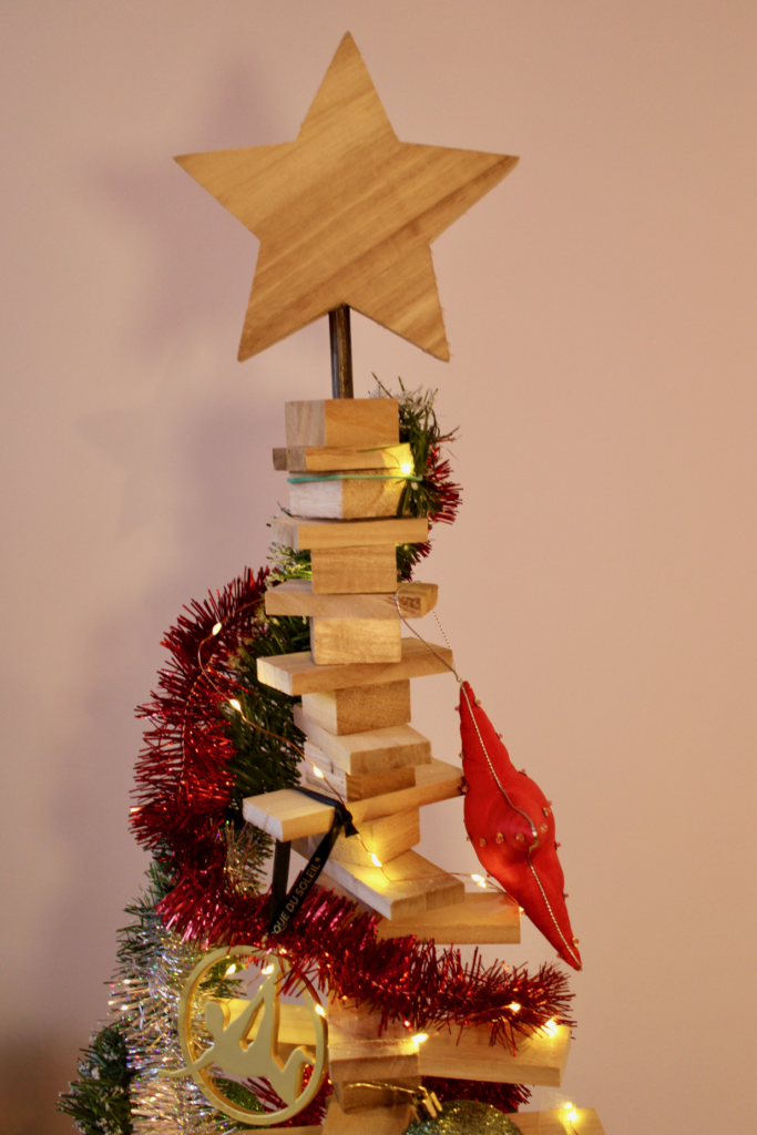 A wooden star sits atop a creatively decorated Christmas tree, which features stacked wooden blocks and colorful ornaments. The tree is adorned with tinsel and warm lights, creating a festive atmosphere.