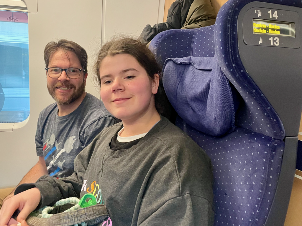 A man and a girl are seated together on a train, both smiling and appearing relaxed. The interior of the train is visible in the background.