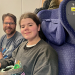 A man and a girl are seated together on a train, both smiling and appearing relaxed. The interior of the train is visible in the background.