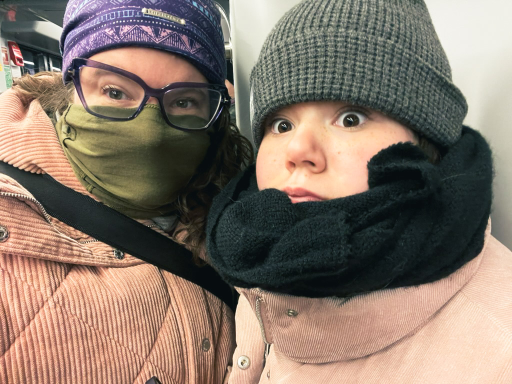 Two women wearing winter clothing and masks are taking a selfie on a subway. They appear surprised or amused, with one woman wearing glasses and a scarf, while the other has a knitted hat and a thick scarf.