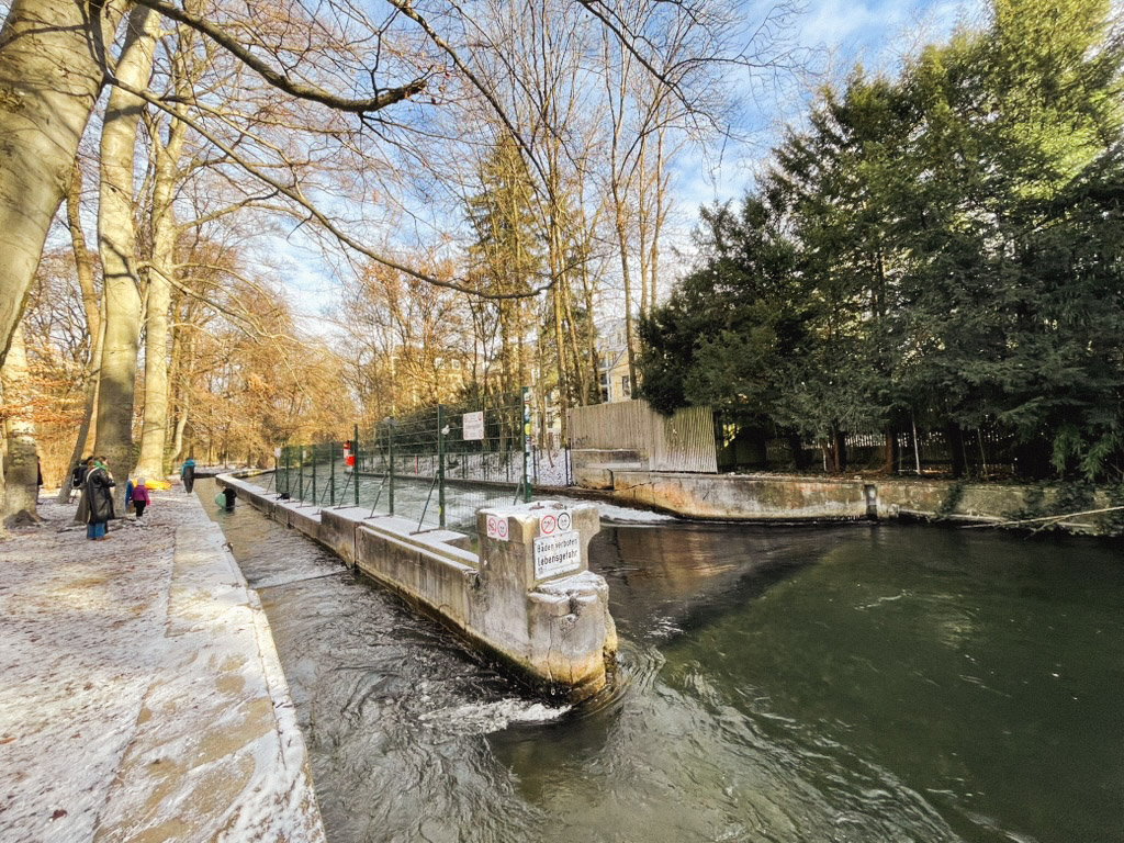 A serene river scene bordered by trees, with a pathway alongside and a small structure extending into the water. The atmosphere suggests a peaceful outdoor setting, ideal for leisurely walks.