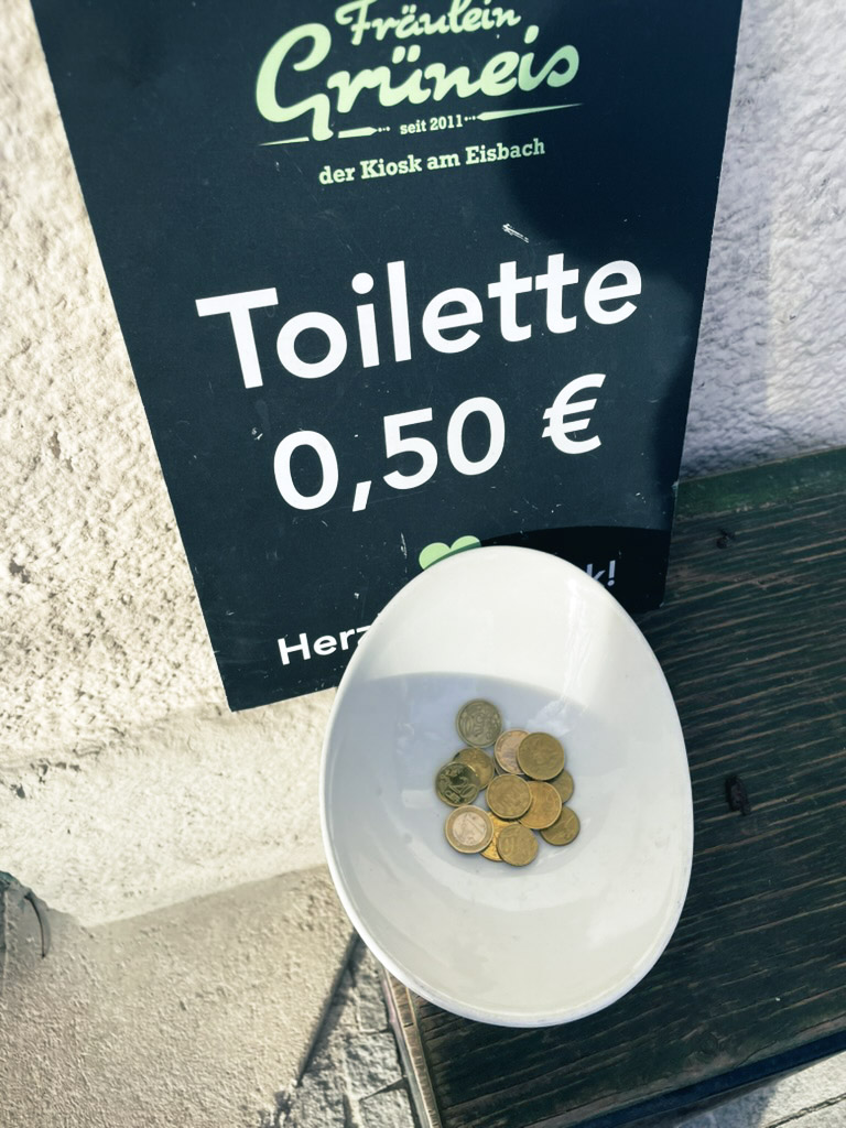 A bowl containing coins sits next to a sign indicating a 0.50 € charge for using the toilet.