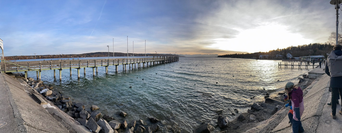 A panoramic view of a pier extending into calm waters, with a sunset illuminating the sky. People are seen along the shore, enjoying the serene landscape.