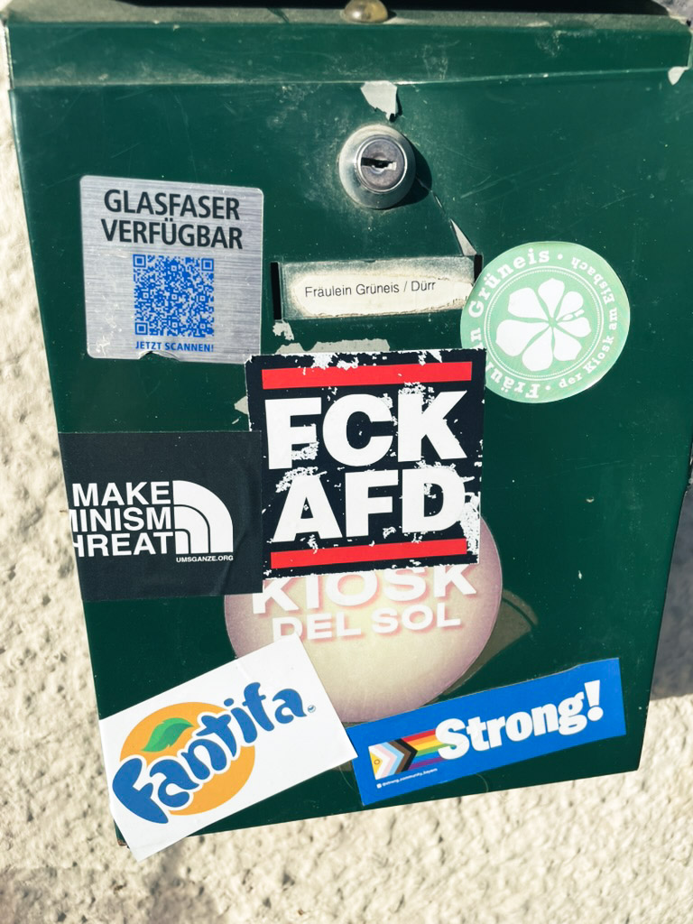 A green utility box covered with various stickers, including one that reads "FCK AFD" and another that says "Fantifa" in the style of the Fanta logo. The box features a slot for access, suggesting it may contain a service or information.