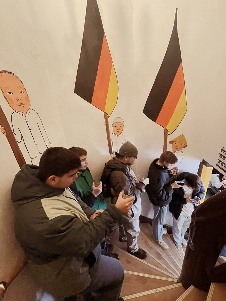 A group of people stands on a staircase, some holding cups of green tea, while colorful murals of people and flags adorn the walls. The atmosphere appears casual and lively.