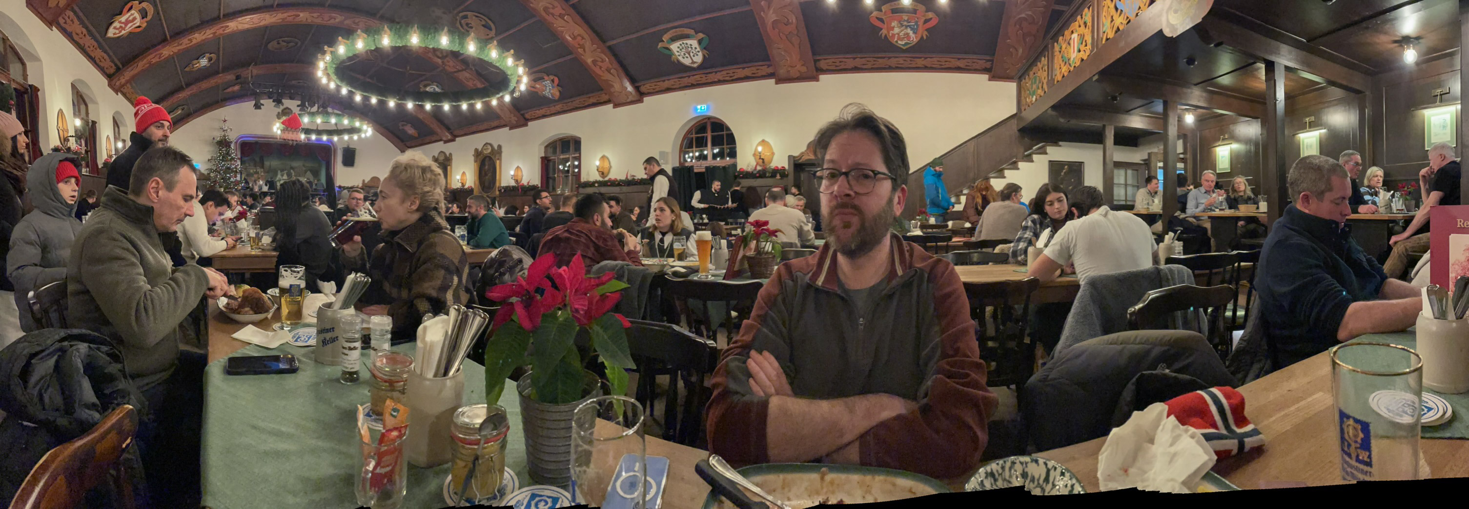 A panoramic view of a bustling restaurant filled with diners, featuring Kristian sitting at a table adorned with flowers and various dishes. The warm lighting and wooden beams create a cozy atmosphere.
