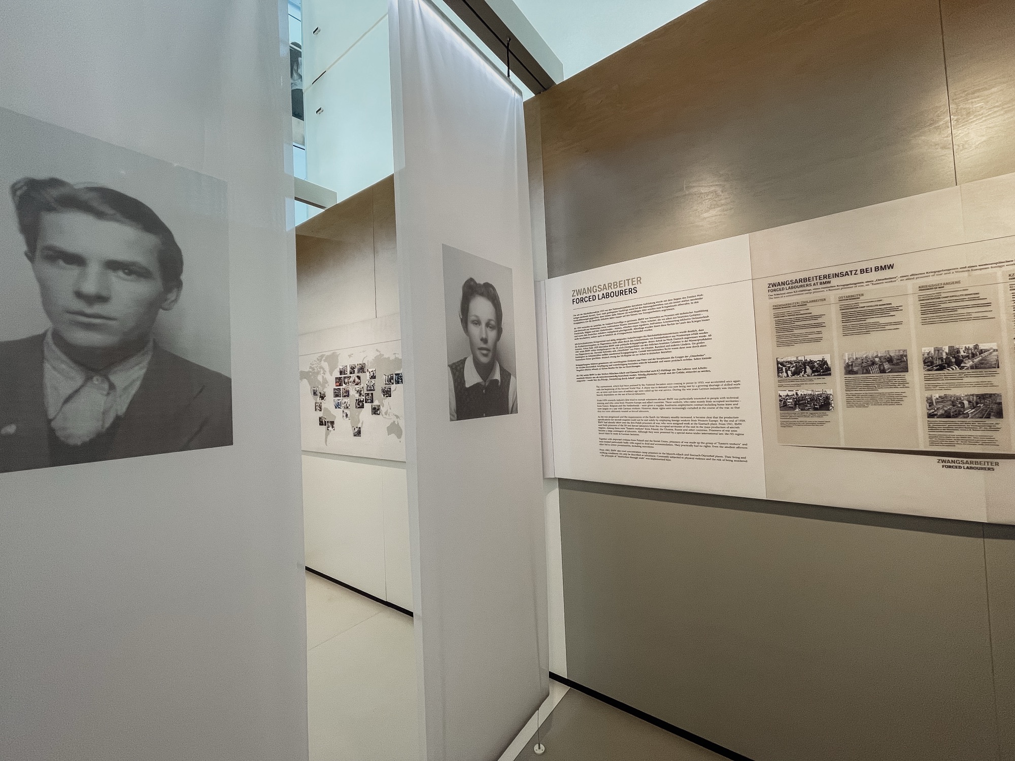 A corridor in an exhibition at the BMW Museum featuring black-and-white portraits of forced laborers on the walls alongside informational panels.