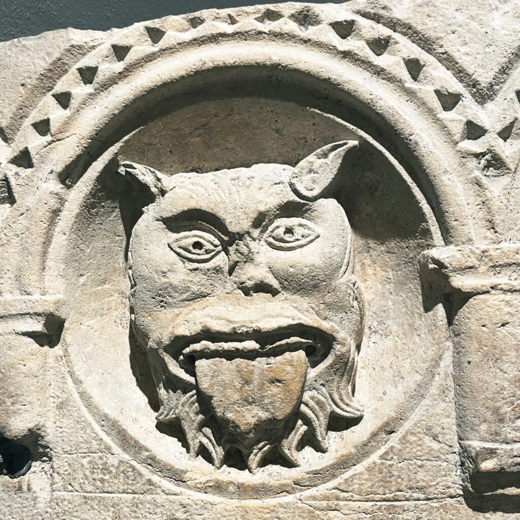 A carved stone relief depicting a grotesque face with pointed ears and a long tongue, framed by an arch with decorative patterns.