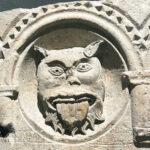 A carved stone relief depicting a grotesque face with pointed ears and a long tongue, framed by an arch with decorative patterns.