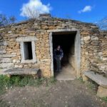 A small ancient stone hut, known as a caborne, with a person standing in the doorway. The structure features a rustic design and is surrounded by greenery under a blue sky.
