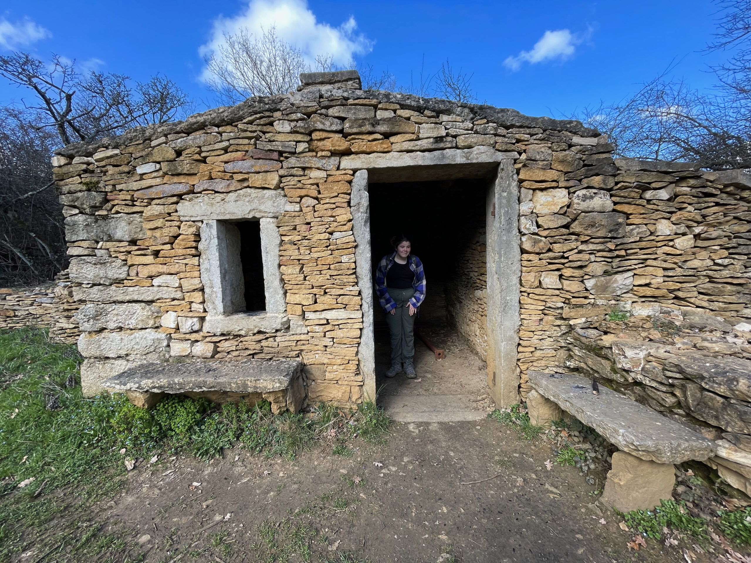 A small ancient stone hut, known as a caborne, with a person standing in the doorway. The structure features a rustic design and is surrounded by greenery under a blue sky.