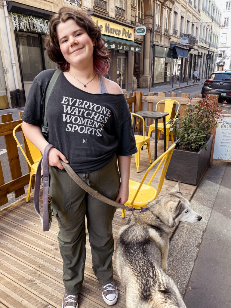 A young person with curly hair stands on a city street, smiling while holding a dog's leash. The background features outdoor seating and buildings typical of an urban area.