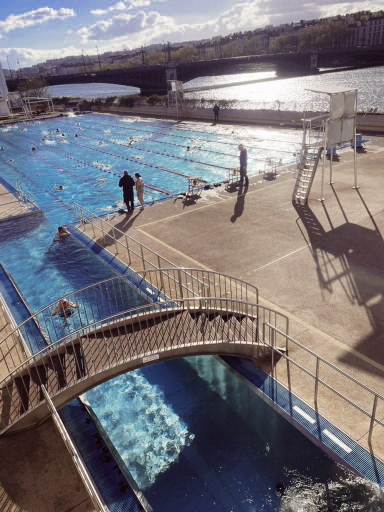Outdoor swimming pool beside the Rhône River, featuring a lap area and a bridge leading to a water slide. The scene includes swimmers enjoying the water under a partly cloudy sky.