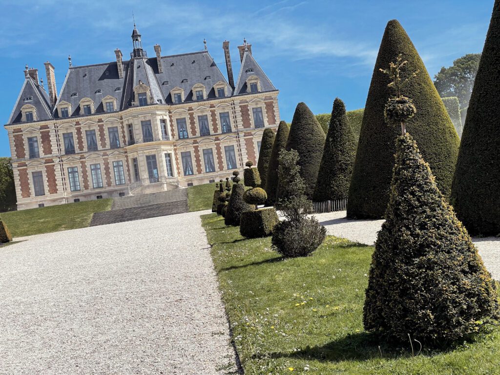 A grand chateau with a blue sky in the background, surrounded by neatly trimmed hedges in a landscaped garden. The scene captures the elegance of the architecture and the artistry of the topiary.