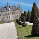 A grand chateau with a blue sky in the background, surrounded by neatly trimmed hedges in a landscaped garden. The scene captures the elegance of the architecture and the artistry of the topiary.