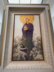 A large modern painting ("Entrust," by Inti) depicts the Virgin Mary holding a child, surrounded by angels and figures, with a radiant halo above her head. The artwork features vibrant colors and intricate details, framed within an ornate border.