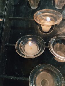 A close-up view of several lit candles in glass holders, with one candle prominently burning in the foreground. The setting appears to be a place of reflection or prayer.