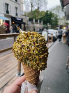A hand holds a cone of Lebanese ice cream topped with crushed pistachios, set against a bustling street scene.