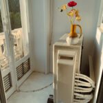 A portable air conditioner sits in a bright room next to a vase with flowers, with large windows providing a view outside. The space is designed to keep cool during a heat wave.