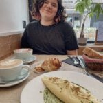 A person with curly hair smiles while sitting at a café table, enjoying a meal that includes an omelet on a plate with green sauce, alongside coffee and pastries.