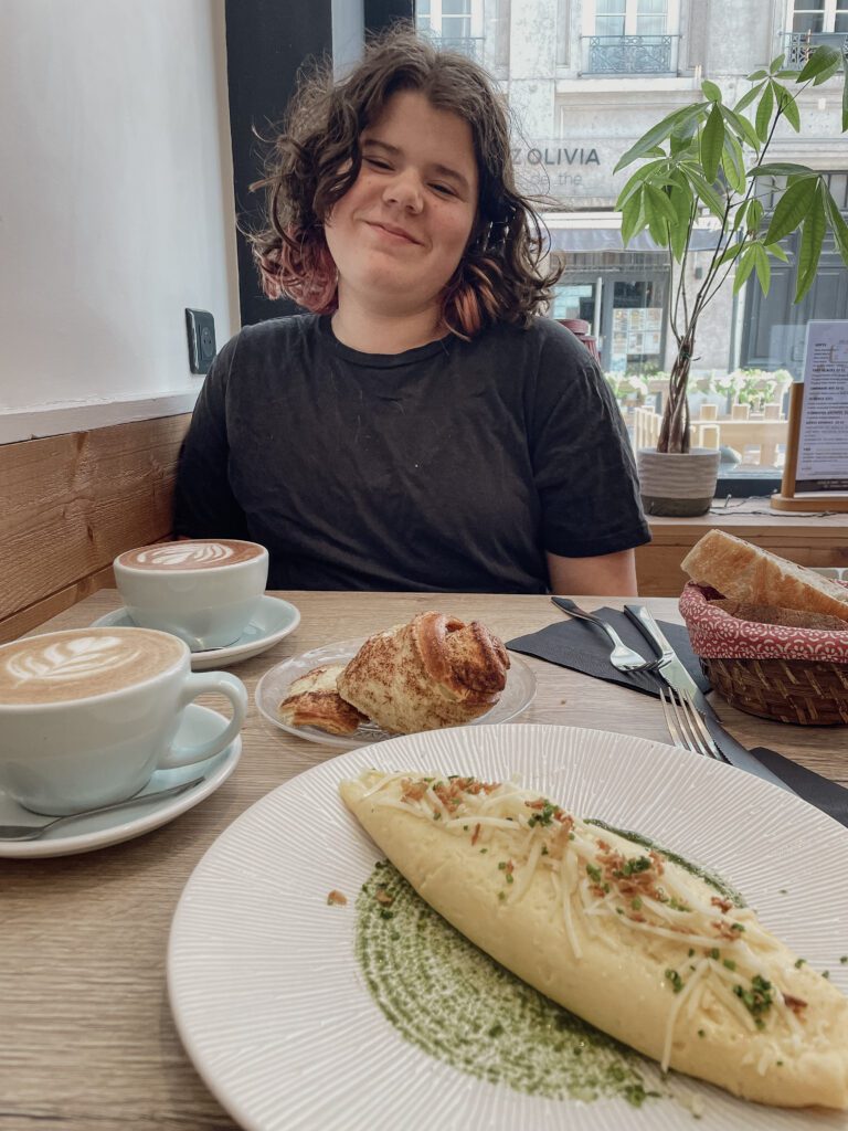 A person with curly hair smiles while sitting at a café table, enjoying a meal that includes an omelet on a plate with green sauce, alongside coffee and pastries.