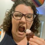 A woman with curly hair and glasses playfully pretends to take a bite of a small cupcake while making a funny face. Colorful decorations are visible in the background.