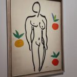 A simple line drawing of a nude figure surrounded by colorful oranges and green leaves, displayed in a wooden frame. The artwork reflects the style of Henri Matisse.