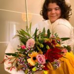 A young person smiles while holding a large bouquet of colorful flowers. The setting appears to be indoors, with a light-colored background.