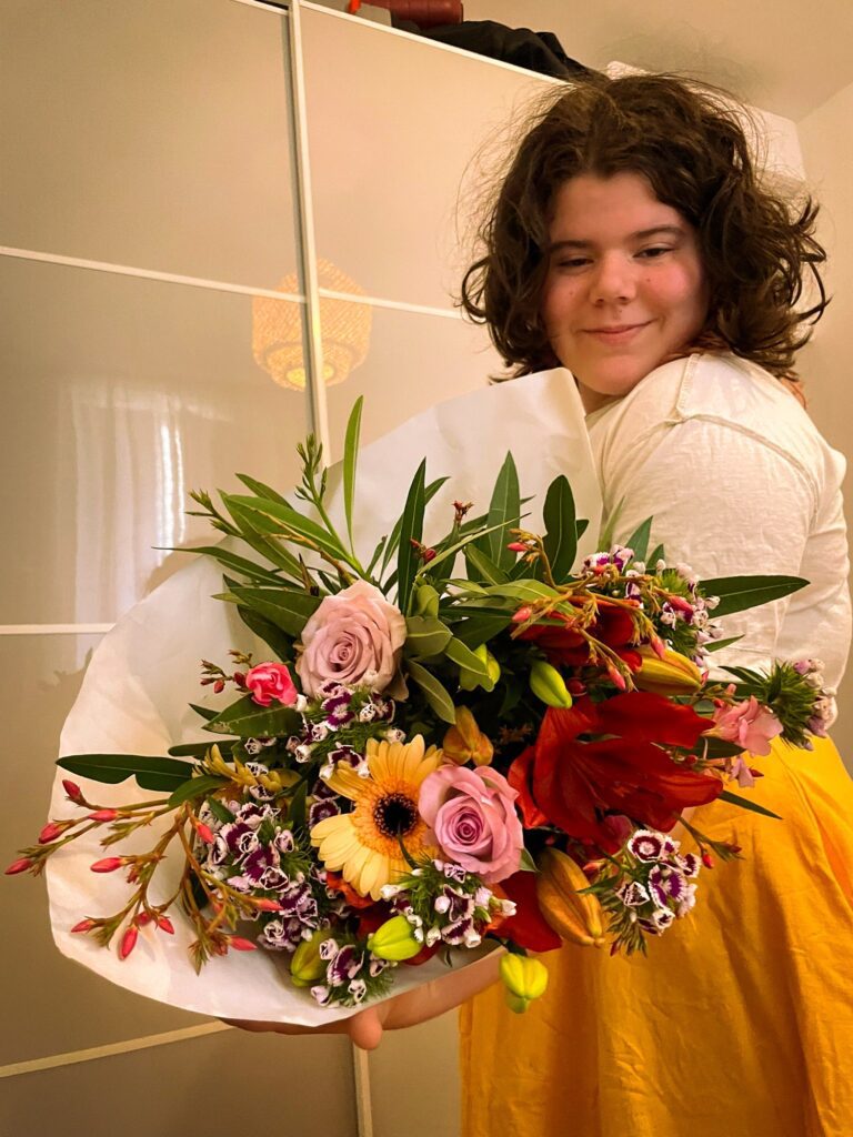 A young person smiles while holding a large bouquet of colorful flowers. The setting appears to be indoors, with a light-colored background.