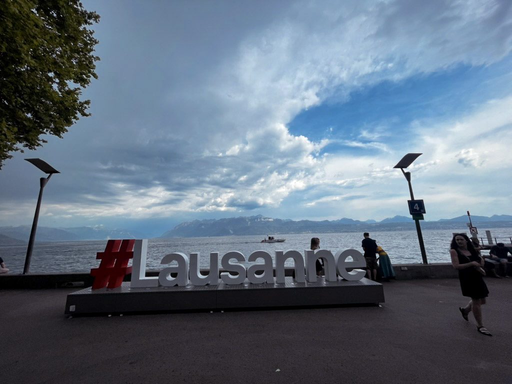 A large "#Lausanne" sign is displayed along the waterfront, with people enjoying the view and a cloudy sky above. The scene captures a vibrant atmosphere near Lac Léman.