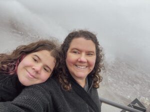 Two smiling women pose together on a foggy day, with a scenic view of a city in the background. They are wearing warm clothing, suggesting a cool climate.