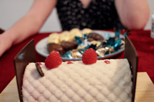 A rectangular cake adorned with raspberries sits on a wooden board, with a plate of assorted chocolates and treats in the background. The setting features a red tablecloth, creating a festive atmosphere.