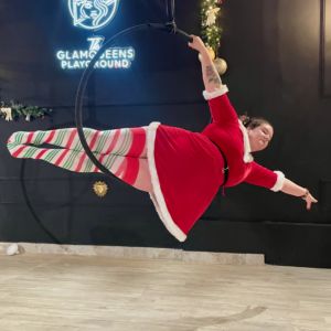 Hope in a festive red costume poses gracefully in an aerial hoop, showcasing a playful holiday spirit. The backdrop features Christmas decorations, enhancing the cheerful atmosphere.