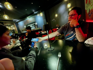 A child is handing a smartphone to an adult at a dimly lit restaurant table, with other diners visible in the background. The atmosphere appears casual and social.