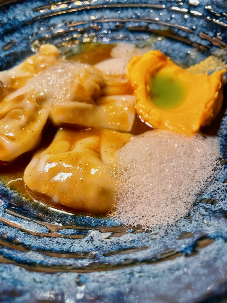 A plate with delicate dumplings served in a flavorful broth, accompanied by a vibrant sauce and a frothy garnish. The dish is presented on a textured blue plate, highlighting its colors and textures.