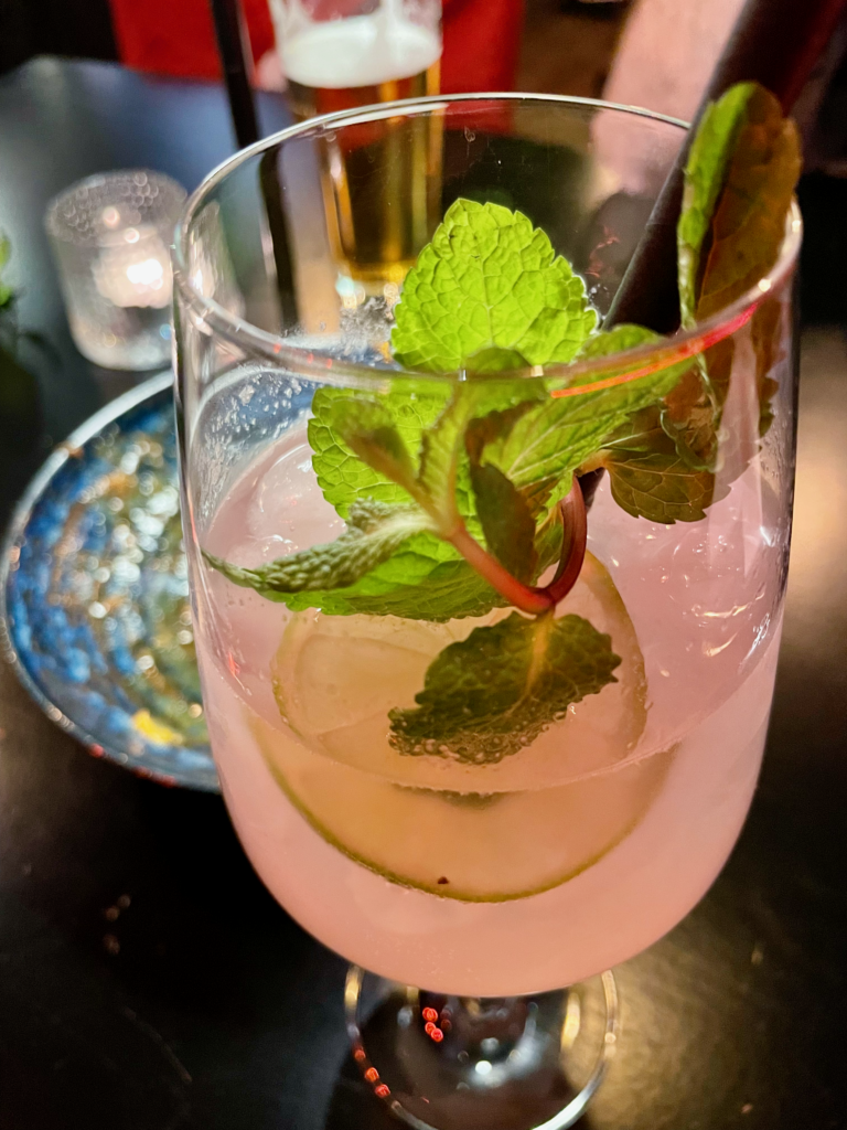 A refreshing cocktail garnished with fresh mint leaves, served in a tall glass with a hint of pink color. The drink is placed on a table with a blurred background of a cozy setting.