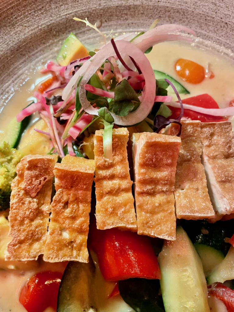 A vibrant dish featuring sliced crispy tofu atop a colorful assortment of fresh vegetables, garnished with herbs and thinly sliced onions. The presentation highlights a mix of textures and flavors.