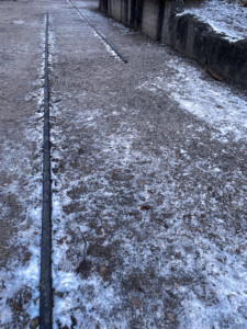 Train tracks outside the Dachau concentration camp. A gravel path with two parallel metal tracks running along it, partially covered in frost. The surrounding area appears cold and unkempt.