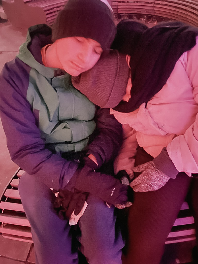 Two people resting closely together on a bench, wearing warm clothing, with their heads leaning against each other. The scene is illuminated with a soft, warm light.