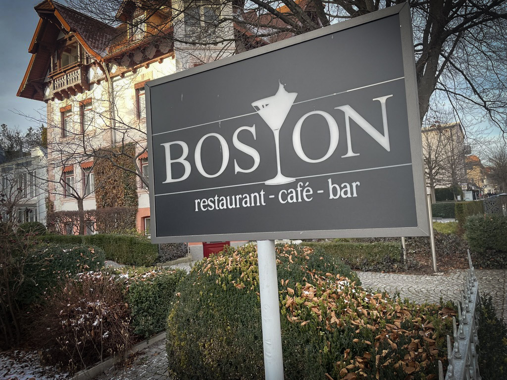 A sign displaying the name and services of the establishment "BOSTON," which offers dining and drinks, is prominently placed outside a building surrounded by greenery.
