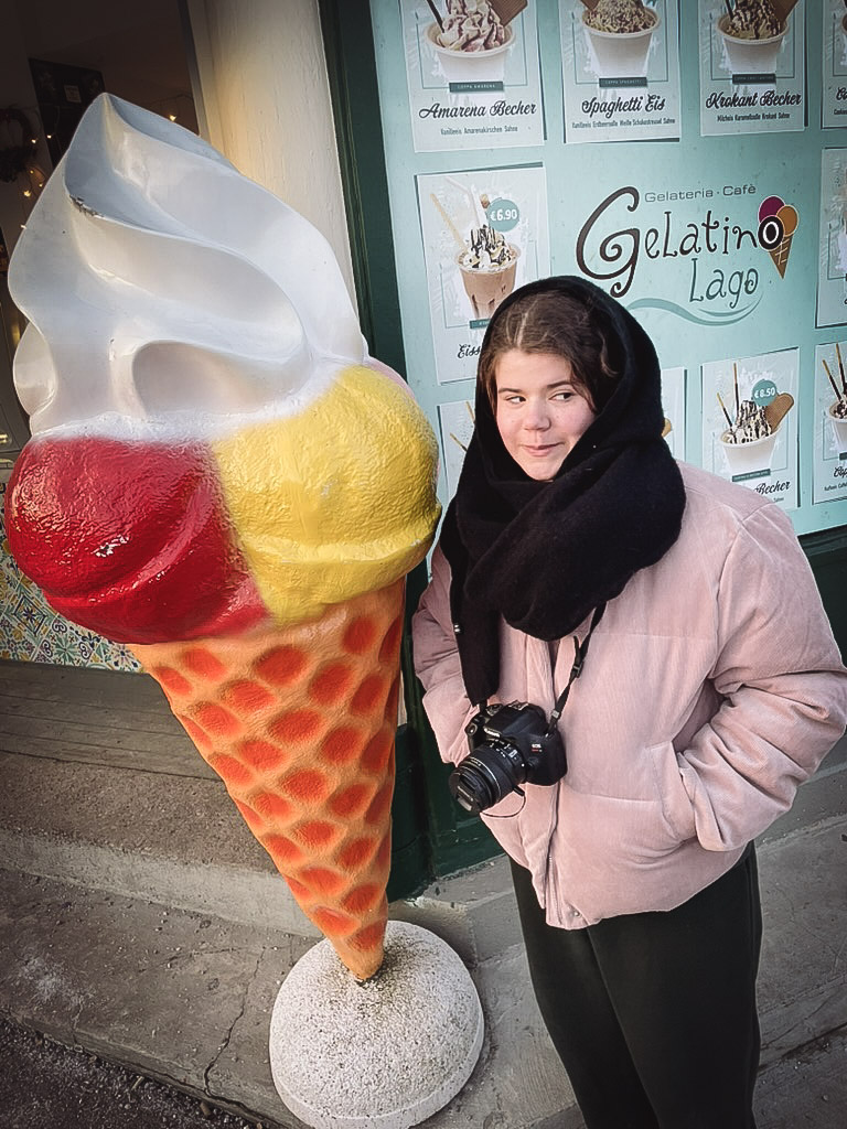 Lilian stands next to a large, colorful ice cream cone sculpture, wearing a warm jacket and scarf, with a camera in her hands. The background features a shop window with various gelato flavors displayed.