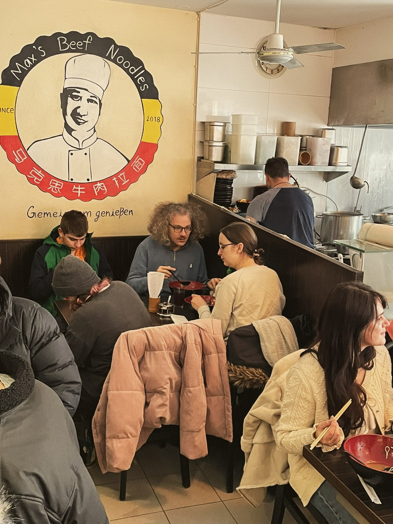 A bustling restaurant interior with patrons enjoying their meals, featuring a prominent mural of a smiling chef on the wall. The atmosphere is lively, showcasing a mix of diners engaged in conversation.