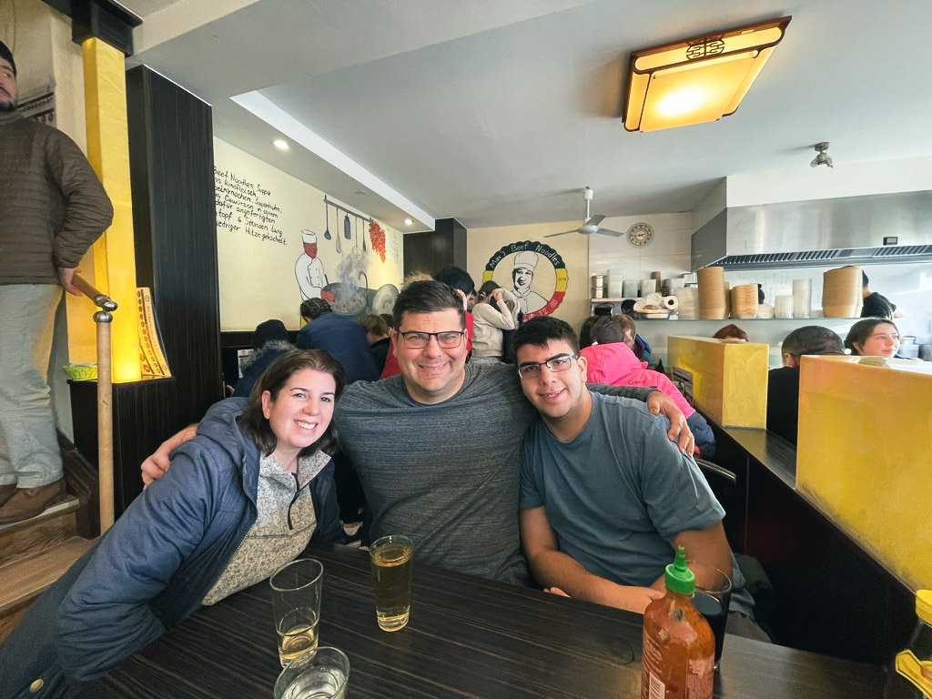 Three friends smiling together at a table in a lively restaurant, with colorful decor and patrons in the background.