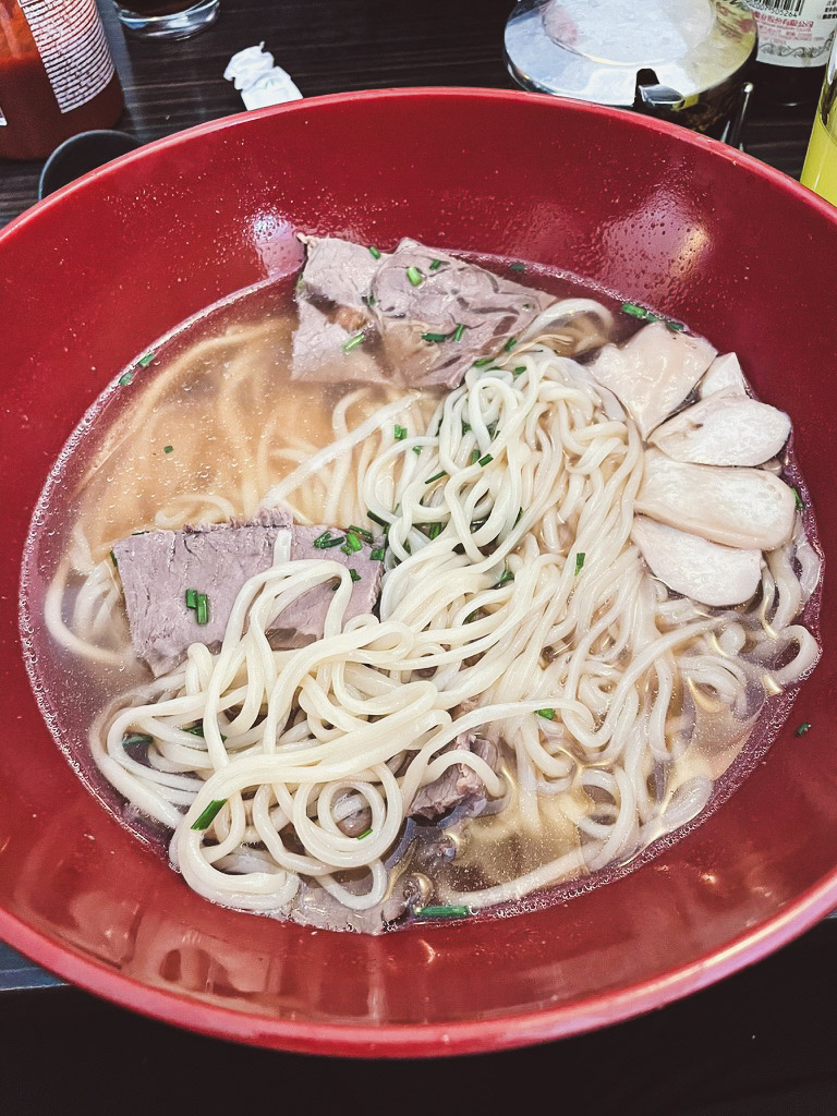 A bowl of ramen featuring noodles, slices of beef, and mushrooms in a rich broth, garnished with green onions.