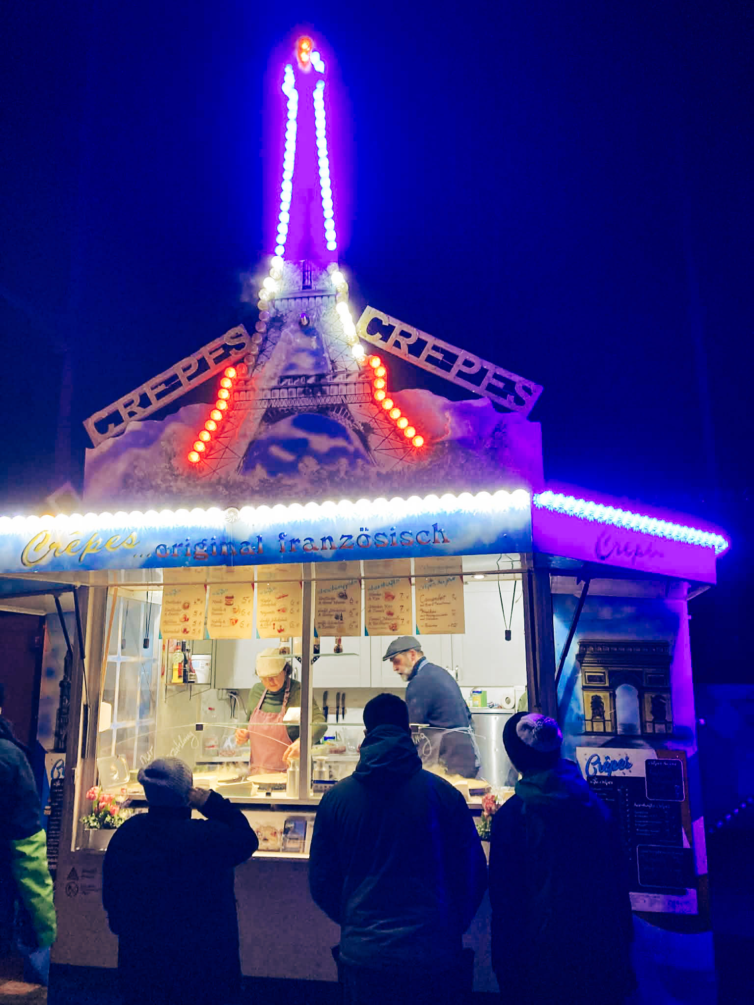 A colorful crepe stand illuminated at night, with customers being served inside. The vibrant lights and festive atmosphere create a lively scene.