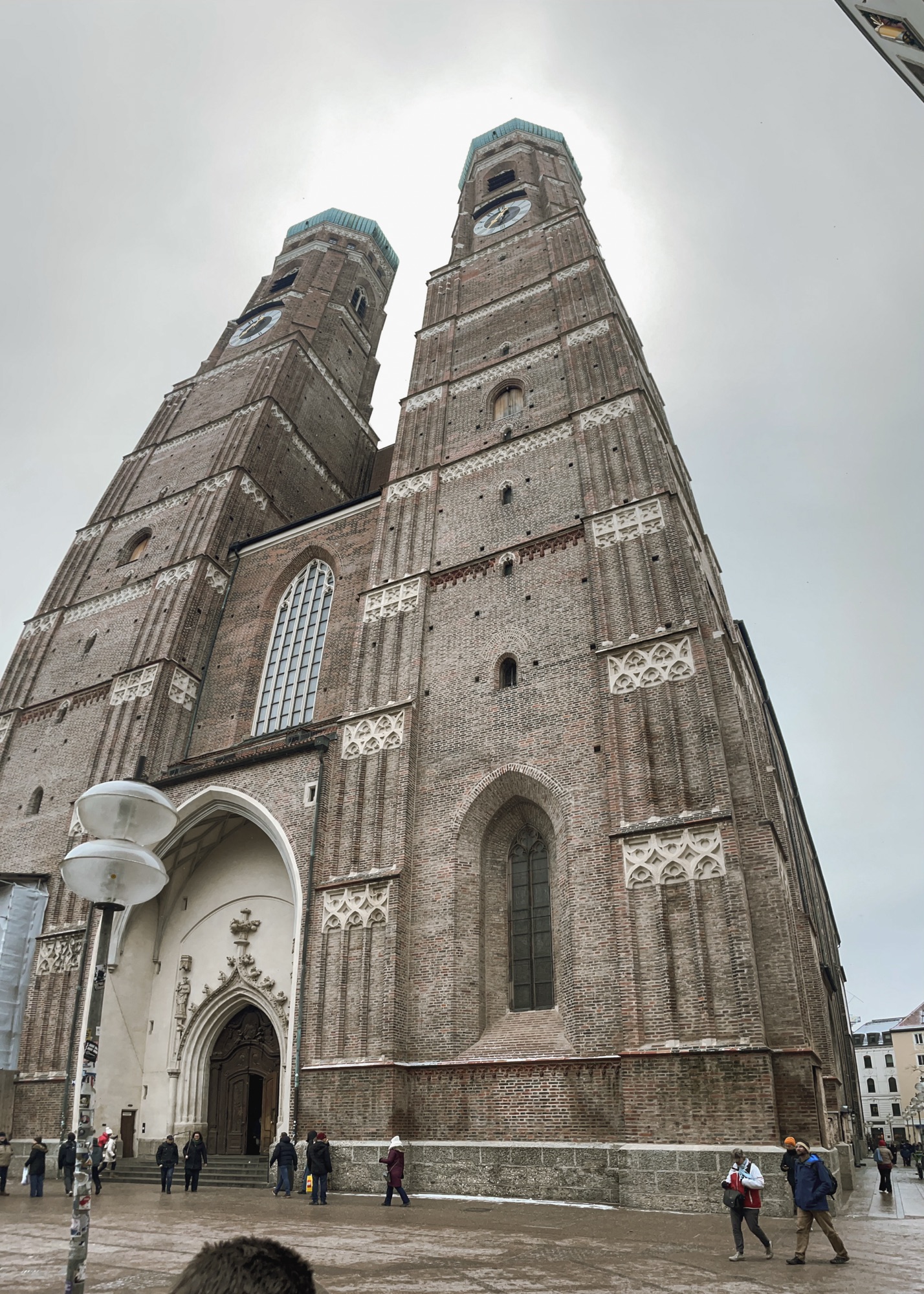 The Frauenkirche, a tall, historic church with two prominent towers stands against a cloudy sky, showcasing intricate brickwork and architectural details. People can be seen walking in the foreground.