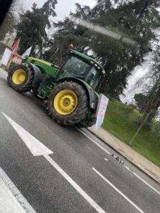 A green tractor is driving down a road, surrounded by protest signs, highlighting farmers' opposition to the EU-Mercosur Trade Agreement.