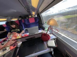 A person works on a laptop while sitting in a train, with a lamp and various items on the table, and a view of the landscape outside the window.